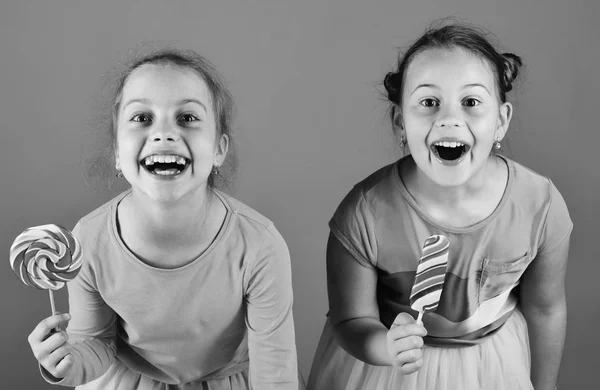 Children with excited faces pose with candies on green background. Sisters with round and long shaped lollipops.