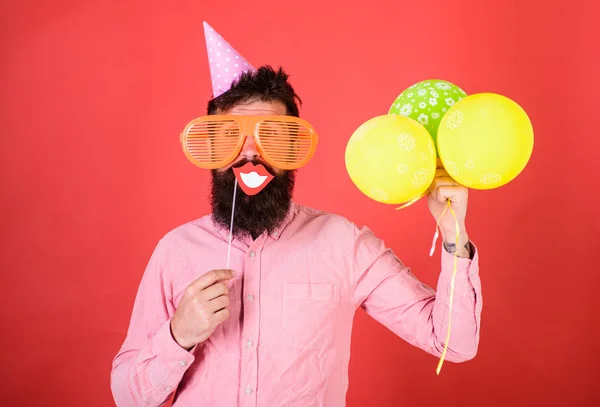Hipster mit riesigen Sonnenbrillen feiern. Mann mit Partyhut und Luftballons feiert. Mann mit Bart auf fröhlichem Gesicht hält lächelnden Mund auf Stock, roter Hintergrund. Konzept für Fotostand-Spaß — Stockfoto