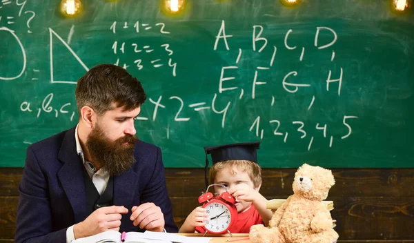 Teacher with beard, father teaches little son in classroom, chalkboard on background. Boy, child on calm face holds alarm clock while teacher talk to kid. Individual lesson concept