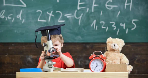 Kid boy i akademiska cap arbete med mikroskopet i klassrummet, svarta tavlan på bakgrund. Barn på upptagen ansikte nära mikroskopet. Smart unge koncept. Första f.d. intresserad av att studera, lärande, utbildning — Stockfoto