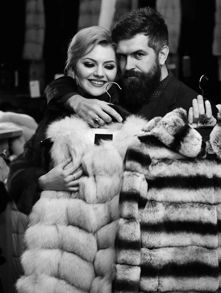 Man and girl with smiling faces hold furry coats on clothes rack background. Guy with beard and woman buy furry coat