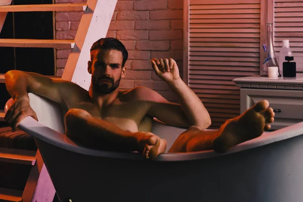 Le sexe et l'érotisme concept. Homme avec barbe et visage inquiet. Macho assis nu dans la baignoire avec des lumières rouges allumées. Guy avec visage concerné dans la salle de bain avec escalier sur le fond — Photo