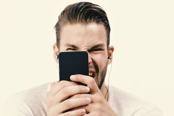 Человек со стильными волосами и разъяренным лицом держит мобильный телефон. Концепция музыки и гаджетов. Мачо в наушниках смотрит видео. Парень с бородой держит MP3 — стоковое фото