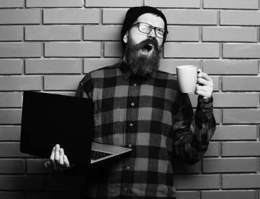 Sakallı, beyaz tenli hippi elinde bardakla dizüstü bilgisayar tutuyor.