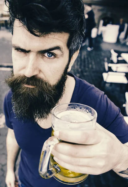 Guy having rest with cold draught beer. Man with beard and mustache holds glass with beer while relaxing at cafe terrace. Hipster on strict face drinking beer outdoor. Draught beer concept