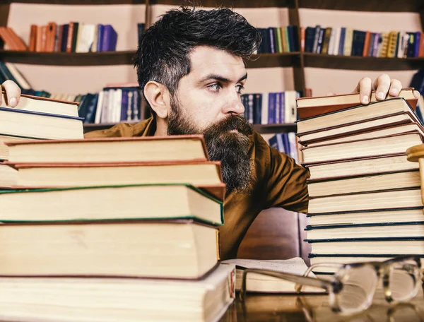 Professor ou estudante com barba senta-se à mesa com óculos, desfocado. Conceito bibliófilo. Homem no rosto estrito entre pilhas de livros, enquanto estudava na biblioteca, estantes de livros no fundo — Fotografia de Stock