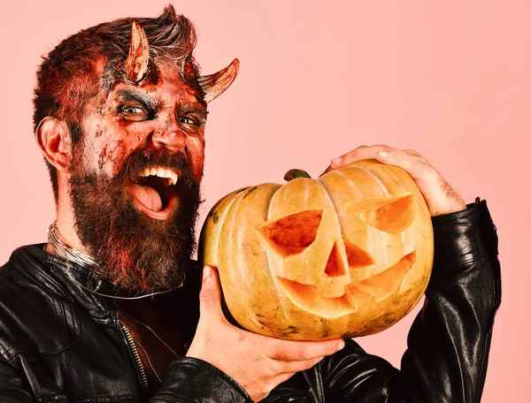 Man wearing scary makeup holds pumpkin on pink background.