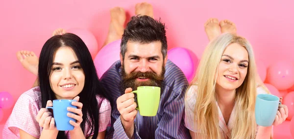 Drei entspannen am Morgen mit Kaffee. Liebhaber-Konzept. Mann und Frau, Freunde auf lächelnden Gesichtern, rosa Hintergrund. Männer und Frauen in Hauskleidung, Schlafanzug. Verliebte trinken Kaffee im Bett — Stockfoto