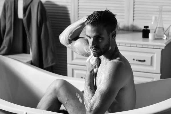 Macho sentado desnudo en la bañera lavándose con esponja y mostrando músculos. Concepto de sexualidad y salud. Hombre con barba y mirada confiada tomando baño —  Fotos de Stock