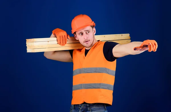 Carpintero, carpintero, fuerte constructor en la cara ocupada lleva la viga de madera en el hombro. Concepto de materiales de madera. Hombre en casco, sombrero duro y guantes protectores apuntando dirección, fondo azul —  Fotos de Stock