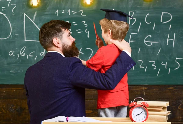 Best friends concept. Teacher with beard, father hugs little son in classroom while discussing, chalkboard on background. Child in graduate cap listening teacher, chalkboard on background, rear view — Stock Photo, Image