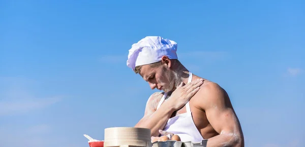 Baker concept. Man on busy face wears cooking hat and apron, sky on background. Chef cook preparing dough for baking with flour. Cook or chef with sexy muscular shoulders and chest covered with flour — Stock Photo, Image
