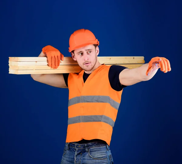 Hombre con casco, sombrero duro y guantes protectores apuntando a la dirección, fondo azul. Carpintero, carpintero, fuerte constructor en la cara reflexiva lleva la viga de madera en el hombro. Concepto de materiales de madera —  Fotos de Stock