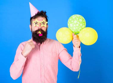 Sakin yüz sakallı adam hava balonları, mavi arka plan tutar. Sürpriz kavramı. Hipster Star gözlük darbeler parti boynuz şeklinde. Tatil öznitelikleri ile parti şapkalı adamı kutluyor