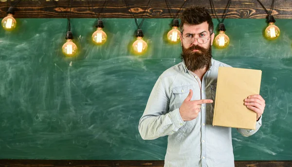 Koncepcja zalecenie. Hipster Brodaty posiada książki, tablica na tle. Nauczyciel w okularach poleca książki. Człowiek z broda i wąsy na stoisku poważne twarz z przodu chalkboard — Zdjęcie stockowe