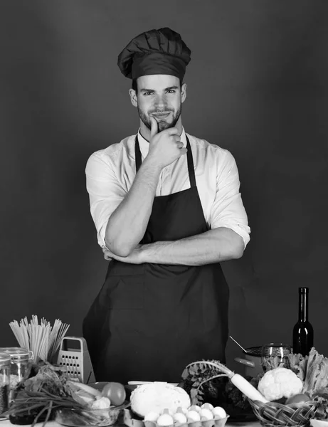 Cook fungerar i köket nära bord med grönsaker och verktyg. Kock med leende ansikte håller handen på hakan, grön bakgrund — Stockfoto