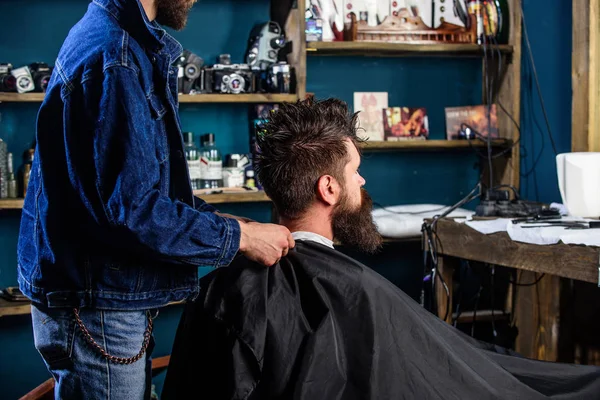 Mann mit Bart und Schnurrbart sitzt im Friseursalon, Schönheitsmittel im Hintergrund. Friseurladen-Konzept. Mann bärtiger Kunde des Hipster-Friseursalons. Hipster mit Bart wartet, während Friseur Hals mit Umhang bedeckt — Stockfoto