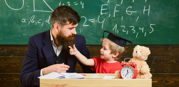 Stygga barn koncept. Far med skägg, lärare lär son, liten pojke. Kid glada distrahera medan du studerar, uppmärksamhetsstörning. Lärare och elev i mortarboard, svarta tavlan på bakgrund — Stockfoto