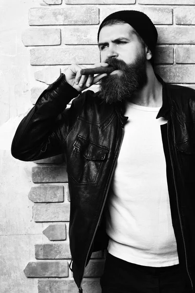 Palenie cygara z czarna skórzana kurtka, kapelusz i białej koszuli na beżowy brick wall studio tło. Brodaty hipster kaukaski brutalny palenie cygar — Zdjęcie stockowe