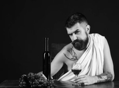 Man with beard holds glass of alcohol on brown background. Sommelier tasting drink. God of wine with tricky face wearing white cloth clipart