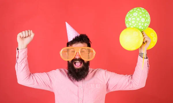 Hipster mit Riesensonnenbrille feiern Geburtstag. Festkonzept. Mann mit Partyhut feiert Feiertag. Mann mit Bart und Schnurrbart auf glücklichem Gesicht hält Bündel Luftballons, roter Hintergrund — Stockfoto