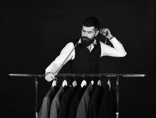 Business fashion and sewing concept, Tailor with serious face holds measuring tape near custom jackets on black background. Designer works near clothes hangers. Man with beard in vest by clothes rack.