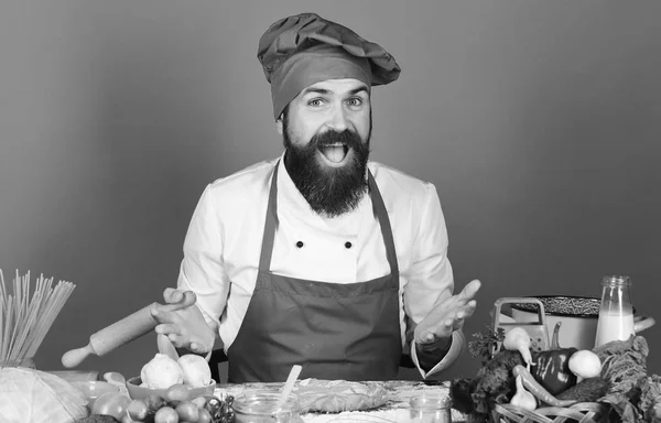 Cuisiner avec le visage joyeux en uniforme bordeaux tient rouleau à pâtisserie, Homme avec barbe est assis par comptoir sur fond bleu . — Photo