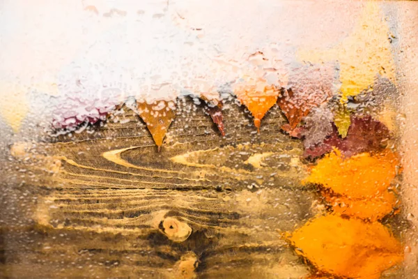 Fondo de otoño, vista a través de vidrio húmedo sobre madera y hojas caídas. Concepto de textura de fondo. Gotas de agua o lluvia sobre vidrio transparente y hojas naranjas de otoño sobre textura de madera — Foto de Stock