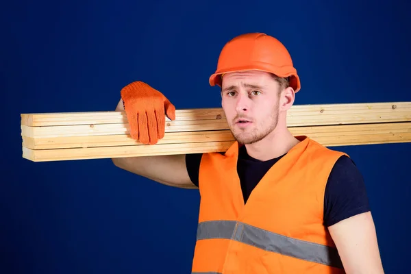 Man in helmet, hard hat and protective gloves holds wooden beam, blue background. Wooden materials concept. Carpenter, woodworker, strong builder on thoughtful face carries wooden beam on shoulder — Stock Photo, Image