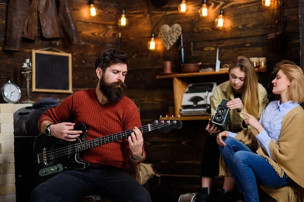 Man with long beard enjoying music. Musician with calm, concentrated face playing for his wife and daughter. Mom helping teenage girl to deal with retro camera. Bearded man tuning electrical guitar