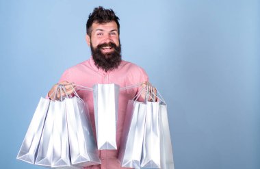Sale and discount concept. Hipster on smiling face shopping addicted or shopaholic. Man with beard and mustache carry shopping bags, light blue background. Guy shopping on sales season with discounts clipart