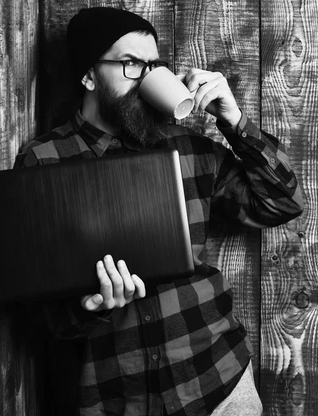 Bearded brutal caucasian hipster holding laptop with cup — Stock Photo, Image