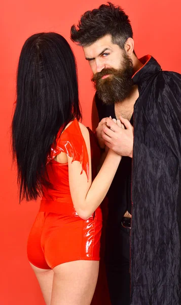 Halloween concept. Couple on strict face play role game. Vampire in cloak and sexy devil girl holds hands. Couple in love, perfect match. Man and woman dressed like vampire, demon, red background — Stock Photo, Image