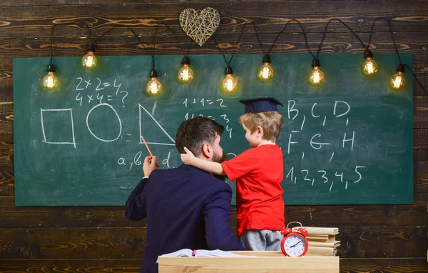 Favoritlärare koncept. Lärare med skägg, far lär lilla son i klassrummet, svarta tavlan på bakgrund. Pojke, barn i graduate cap lyssnande lärare, svarta tavlan på bakgrund, bakifrån — Stockfoto