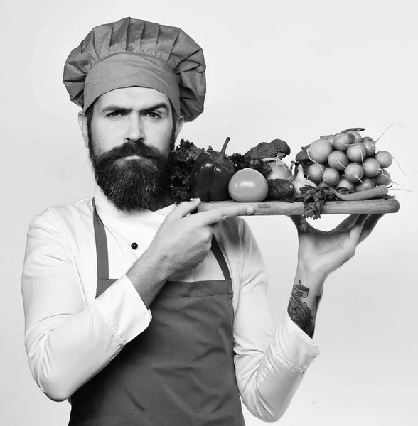 Cook with serious face in burgundy uniform holds salad ingredients. Cooking and vegetarian diet concept. Man with beard