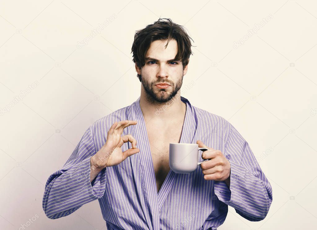 Guy or sportsman in home or bath clothes drinks tea or coffee. Sexy macho in bathrobe with mug showing perfect sign.