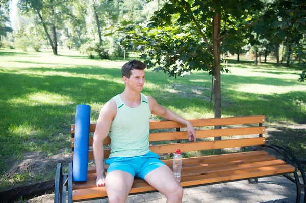 Se acabó el entrenamiento. Hombre con esterilla de yoga y botella de agua sentarse en el banco en el parque. Únete a la práctica de yoga al aire libre. Atleta con equipo de yoga relajante en el parque. El hombre se preocupa por la salud, eligió el yoga al aire libre —  Fotos de Stock