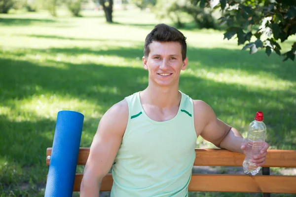 Kierunku zdrowszego stylu życia. Człowiek twarz uśmiechający się matę do jogi i butelka wody siedzieć na ławce w parku. Dołącz do na zewnątrz praktyki jogi. Sportowiec z jogi sprzęt relaks w parku. Mężczyzna wybrał jogi na świeżym powietrzu — Zdjęcie stockowe