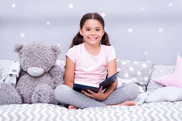 Read fairytale before go to bed. Girl child sit bed with teddy bear read book. Kid prepare to go to bed. Pleasant time in cozy bedroom. Girl kid long hair cute pajamas relax and read book to bear toy