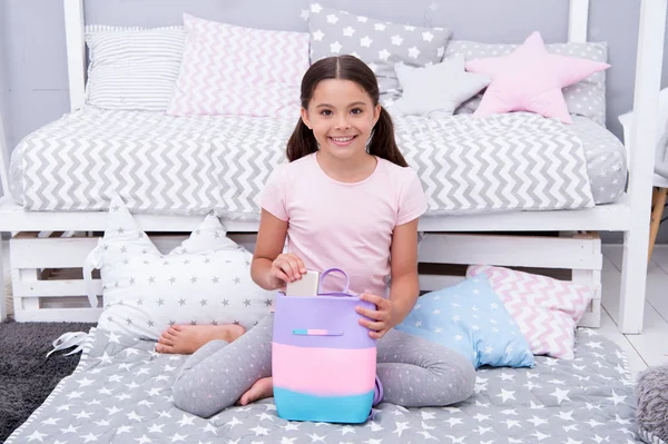 Girlish things. Girl child sit near bed with bag or backpack in her bedroom. Kid prepare to go to bed. Girl kid long hair cute pajamas relaxing in bedroom. Pleasant time to play in cozy bedroom