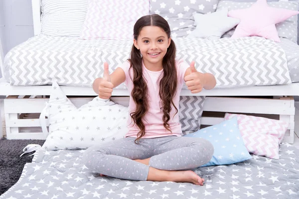 Highly recommend. Girl child sit near bed. Kid prepare to go to bed. Pleasant time in cozy bedroom. Girl kid long hair cute pajamas shows thumbs up gesture. Child satisfied with bedroom interior — Stock Photo, Image