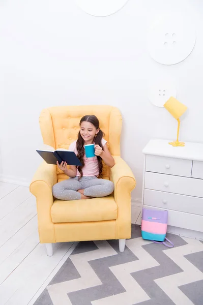 Pleasant evening with book. Girl child sit yellow armchair read book. Kid prepare to go to bed. Pleasant time cozy interior. Girl kid long hair cute pajamas relax and read fairytale book before sleep