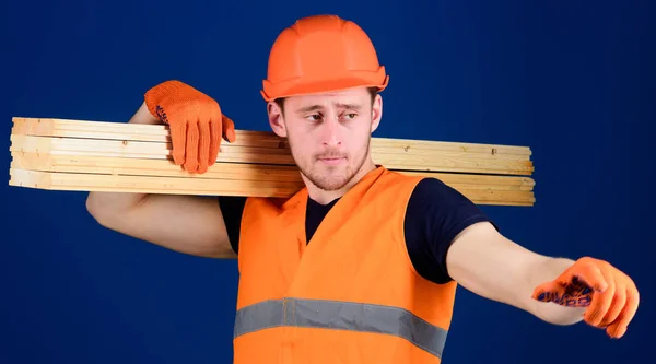 Trä material koncept. Mannen i hjälm, hjälm och skyddshandskar pekar riktning, blå bakgrund. Snickare, möbelsnickare, stark builder på upptagen ansikte bär träbalk på axel — Stockfoto