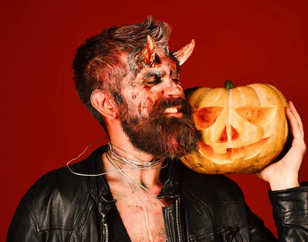 Homme portant un maquillage effrayant tient citrouille sur fond rouge sanglant. Diable ou monstre avec des décorations d'octobre. Démon au visage démoniaque — Photo