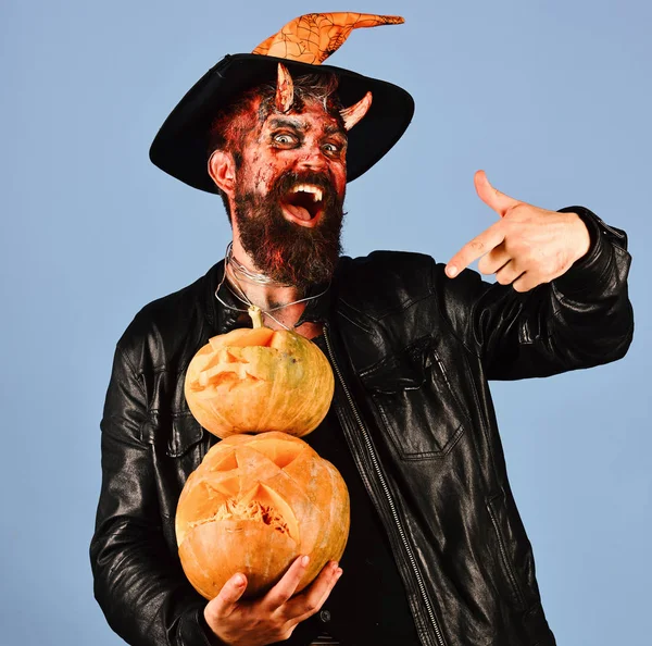 Man wearing scary makeup and witchers hat holds pumpkins on blue background. Monster with October decorations. Halloween spooky party concept.