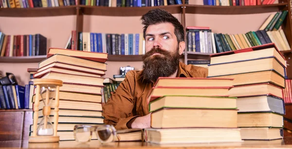 Conceito de bicho-de-biblioteca. Professor ou estudante com barba sentar-se à mesa com ampulheta e óculos, desfocado. Homem no rosto estrito sentar entre pilhas de livros, enquanto estuda na biblioteca, estantes de livros no fundo — Fotografia de Stock