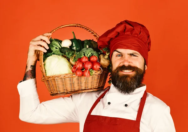 Cook with smile in burgundy uniform holds vegetables basket on shoulder. Chef holds cabbage, radish, broccoli with lettuce and garlic.