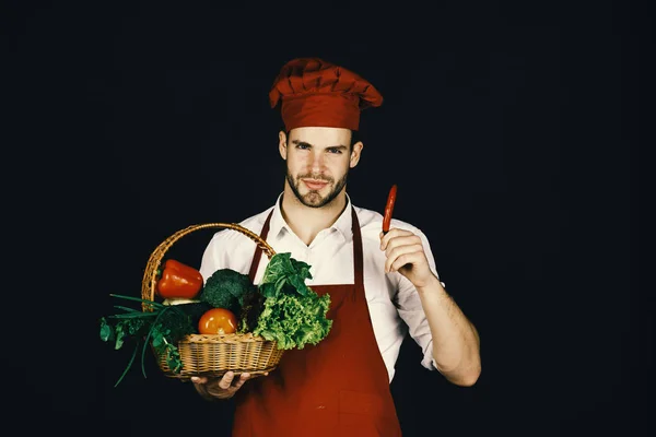 Cook with smiling face, chili and basket of fresh veggies. Chef in burgundy uniform holds red chili in hand. — Stock Photo, Image