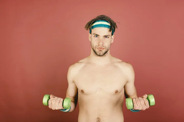 Um tipo com cerdas a usar a cabeça e pulseiras faz exercícios. Homem com tronco nu detém halteres verdes no fundo rosa — Fotografia de Stock