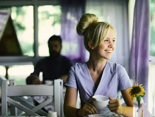 Женщина с улыбающимся лицом пьет горячий напиток. Девушка с светлыми волосами в булочке на светлом фоне кафе — стоковое фото